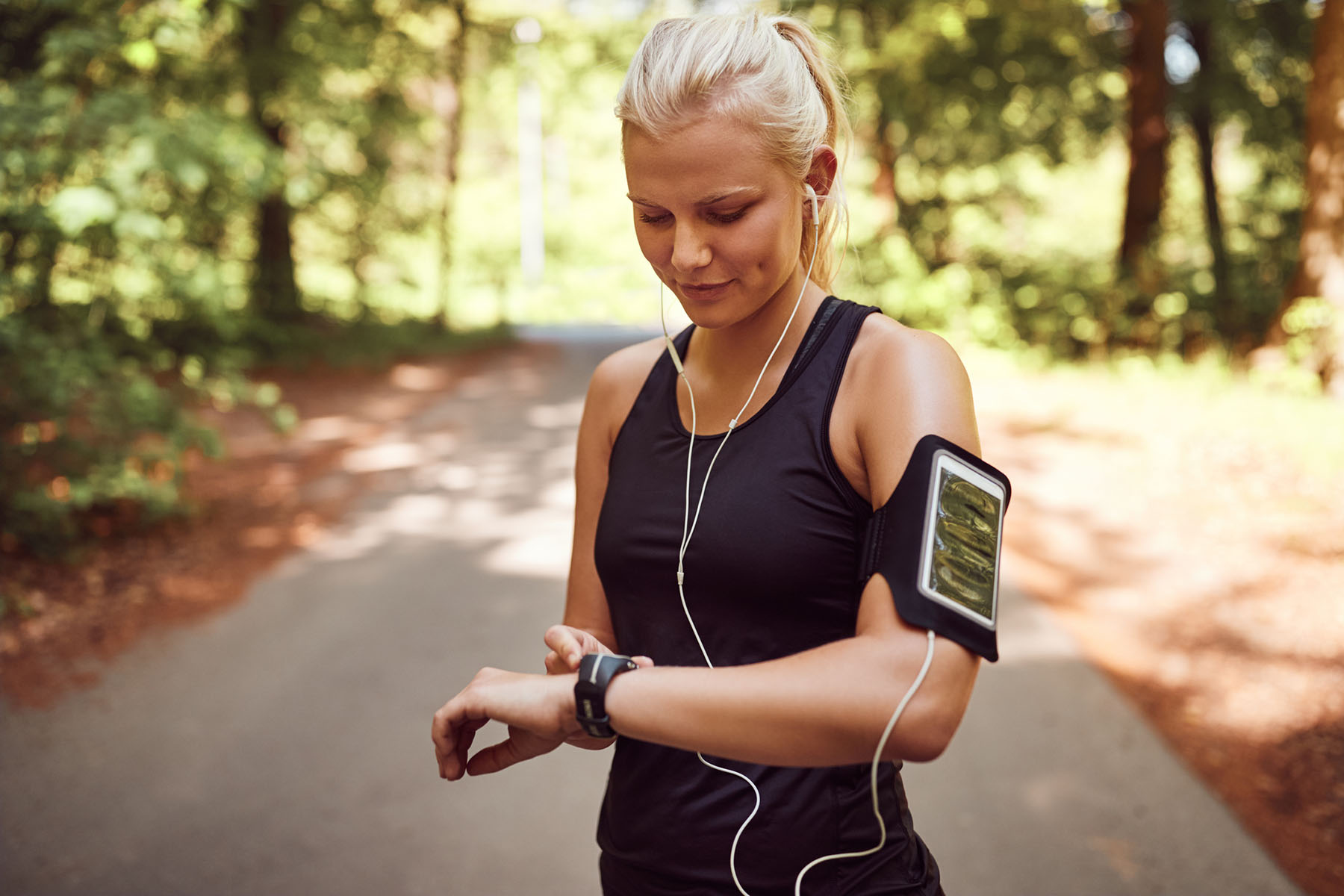 A young woman in athletic wear checking her smartwatch while listening to music with earbuds on a shaded outdoor trail.