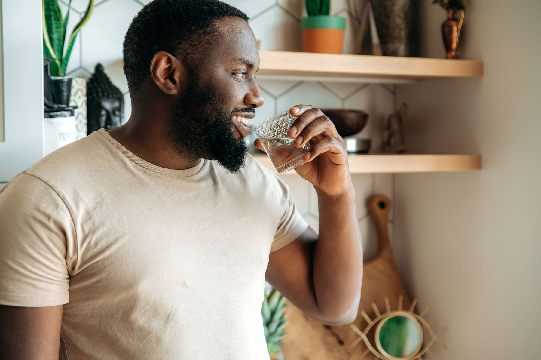 A cheerful man holding a glass of water in a cozy, sunlit kitchen with houseplants and modern decor.
