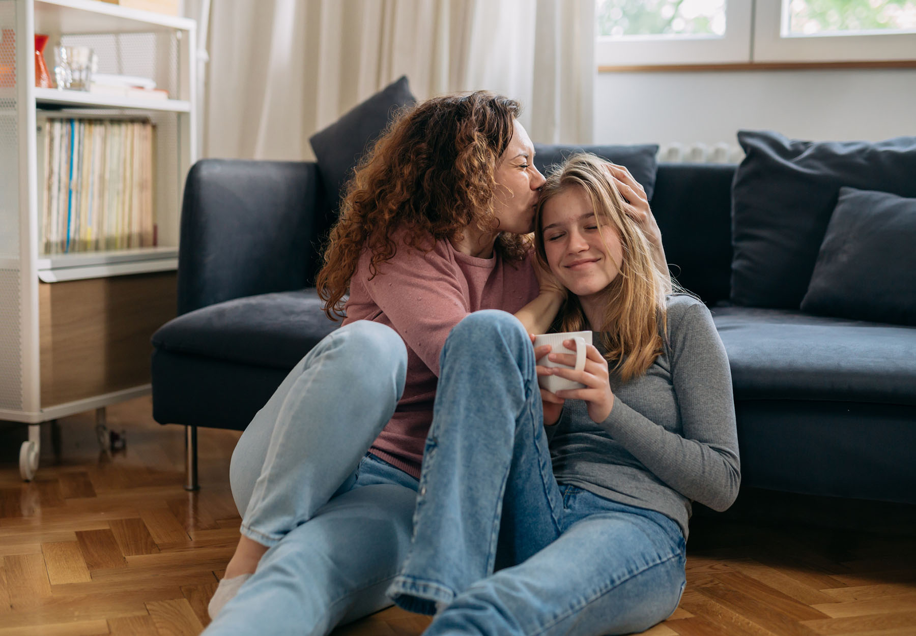 Mother kissing teenage daughter’s forehead on the floor, showing nurturing support and emotional safety through family and adolescent counseling