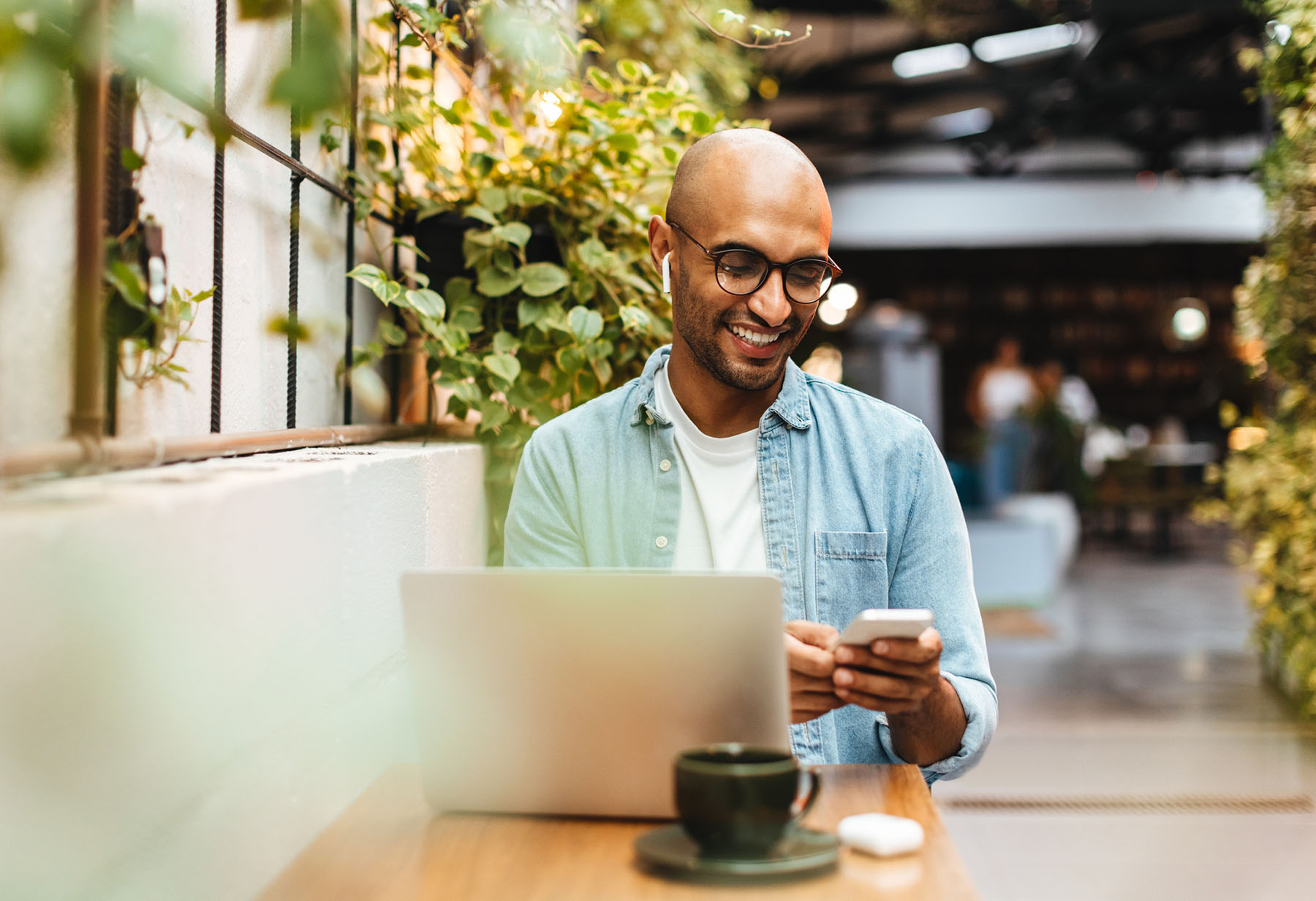 Smiling man working on laptop and phone in cozy cafe setting, representing the flexibility of virtual counseling and telehealth therapy options.
