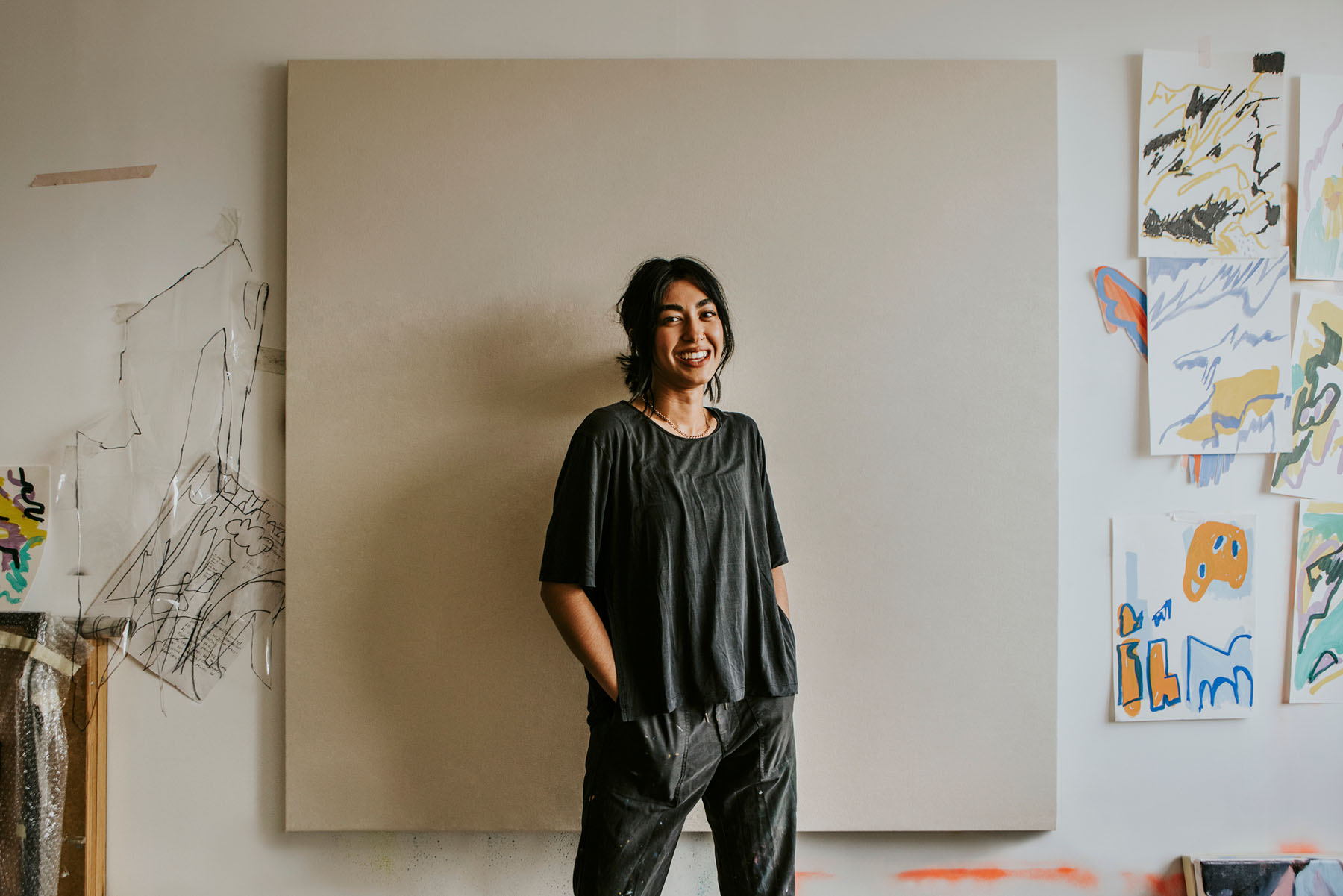 A smiling woman standing with hands in pockets in front of a large blank canvas in an art studio, surrounded by colorful abstract artwork.