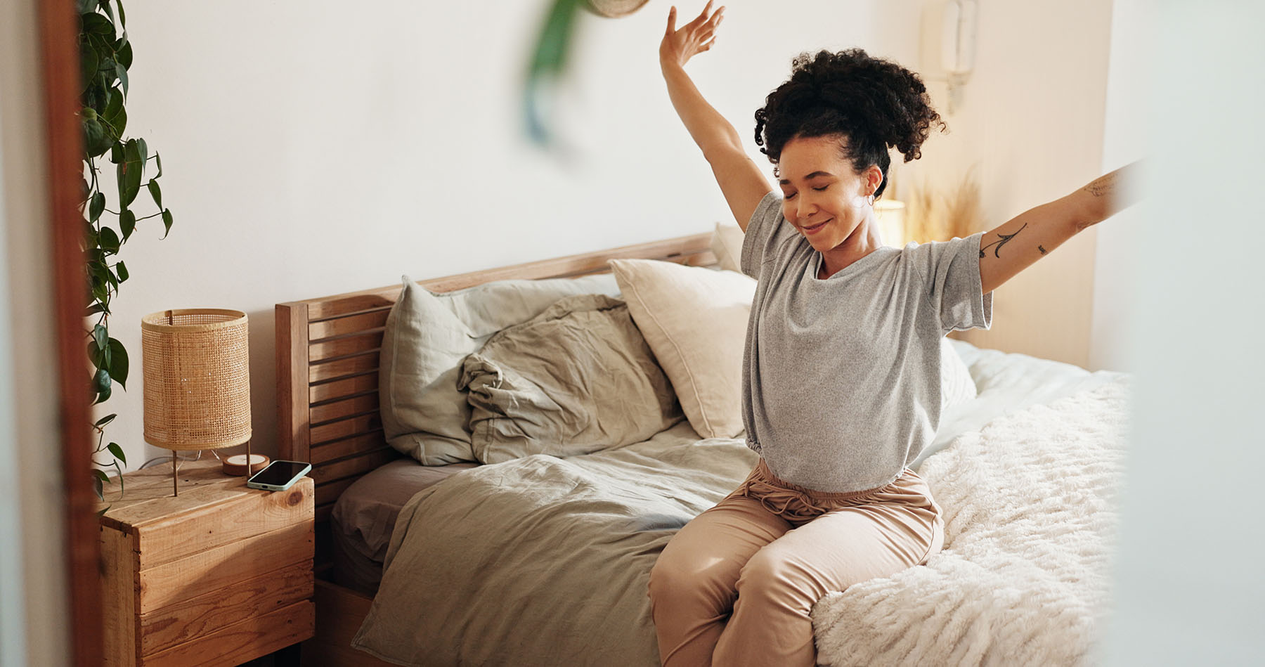 A woman smiling and stretching with arms raised while sitting on the edge of a bed in a softly lit, cozy bedroom.