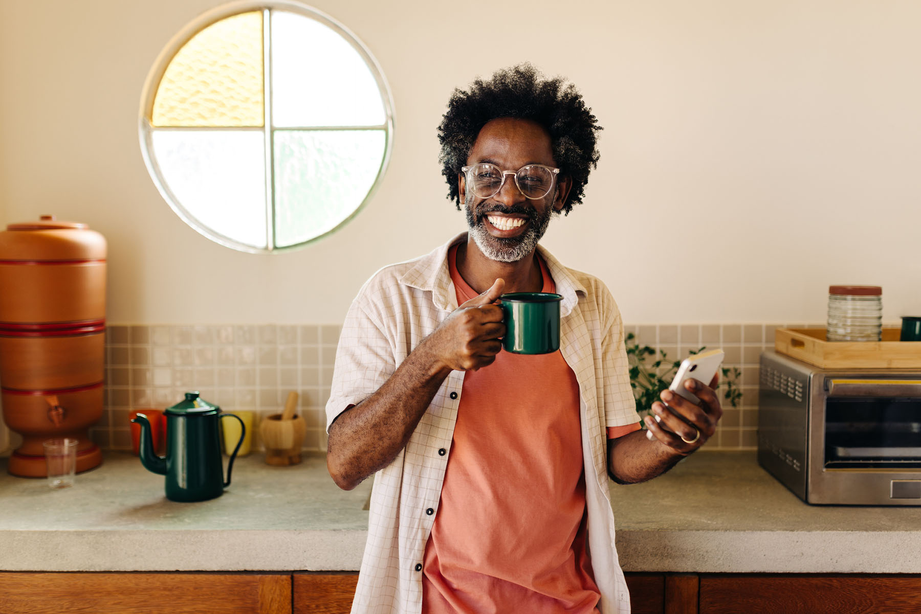 Black man smiling in a bright kitchen, holding a coffee mug and smartphone, representing joy, routine, and the positive impact of mental health support through therapy.