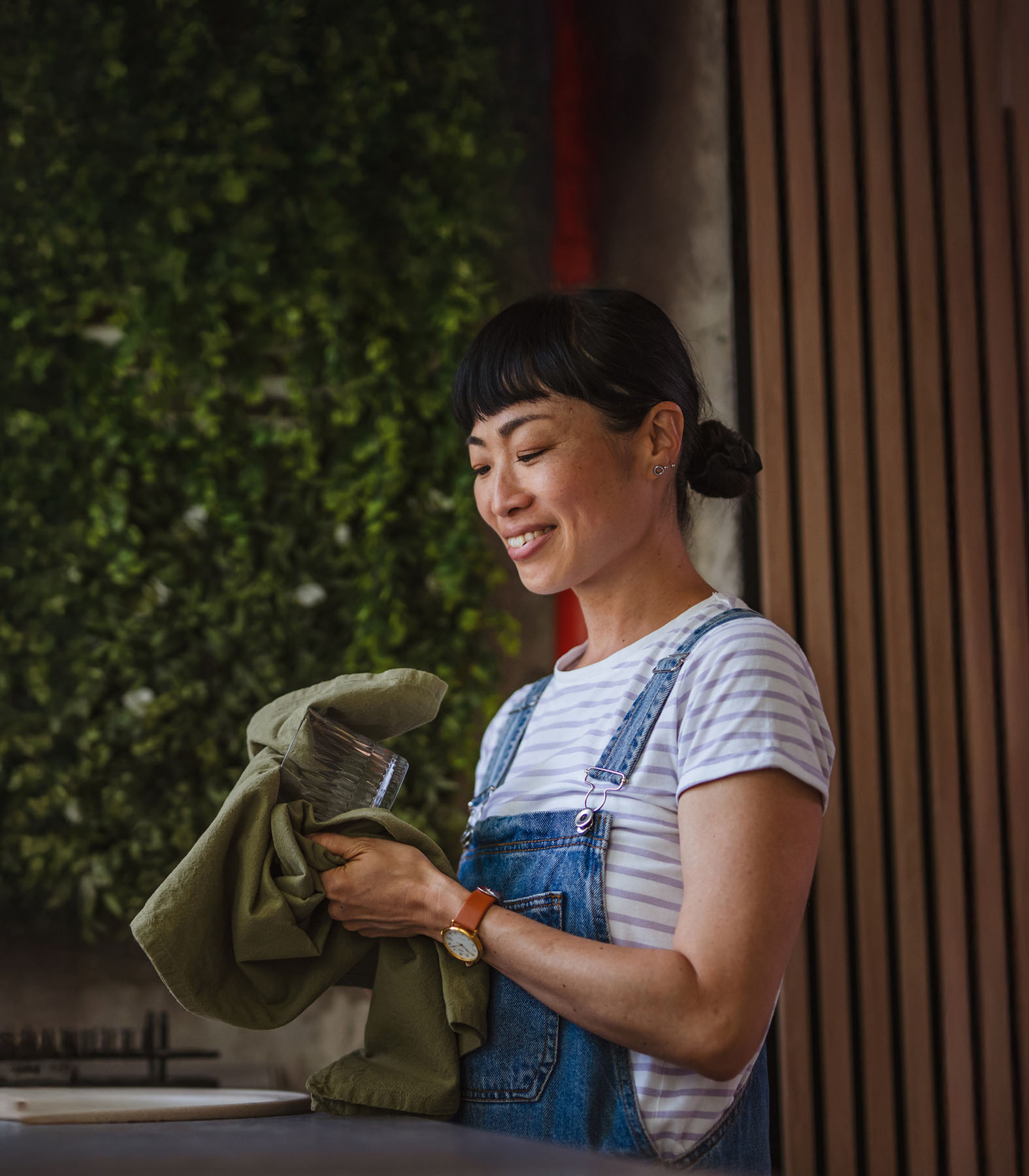 Asian woman smiling while drying dishes in a cozy kitchen setting, representing mindfulness, self-care, and the calming routines encouraged in holistic therapy.
